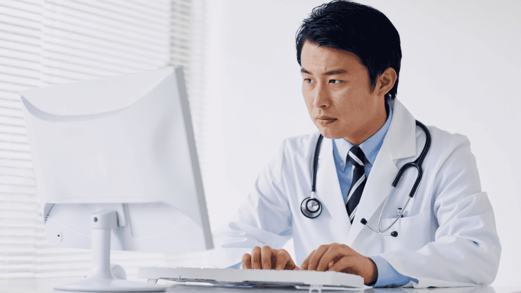 Doctor in a white coat working at a computer, illustrating the challenges of prior authorizations and physician burnout in healthcare.